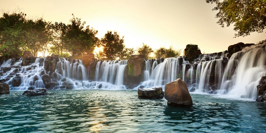 Small seasonal waterfalls surrounded by trees cascade down the green cliffs during sunset.