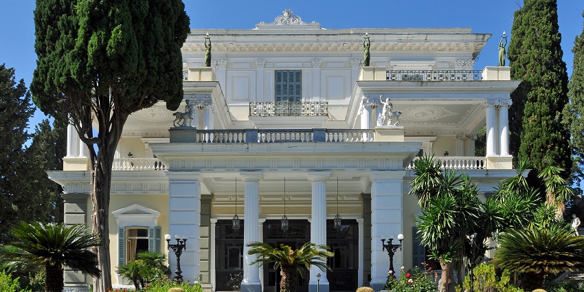 The Neoclassical Achilleion Palace in Corfu, surrounded by lush gardens and tall trees.
