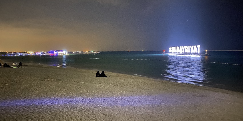 A peaceful night view of Hudayriyat Island beach with illuminated signage reflecting on the calm waters.