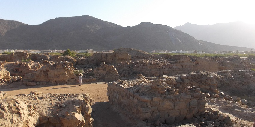 Al-Ukhdood site with ruins, broken stones, a person walking and mountains in the back
