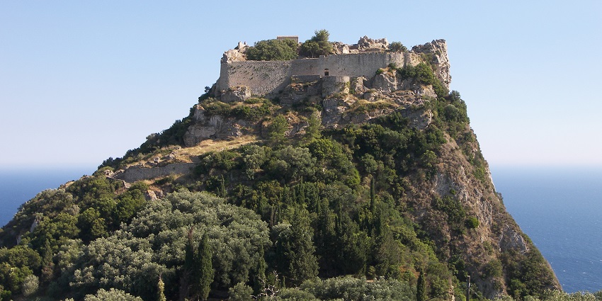 Stone fortress atop a rocky, green hillside under a clear sky, with wooden scaffolding, one of the historical places in Corfu.