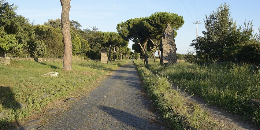 Apian way with small ruined structures on the sides, surrounded by green trees and bushes.
