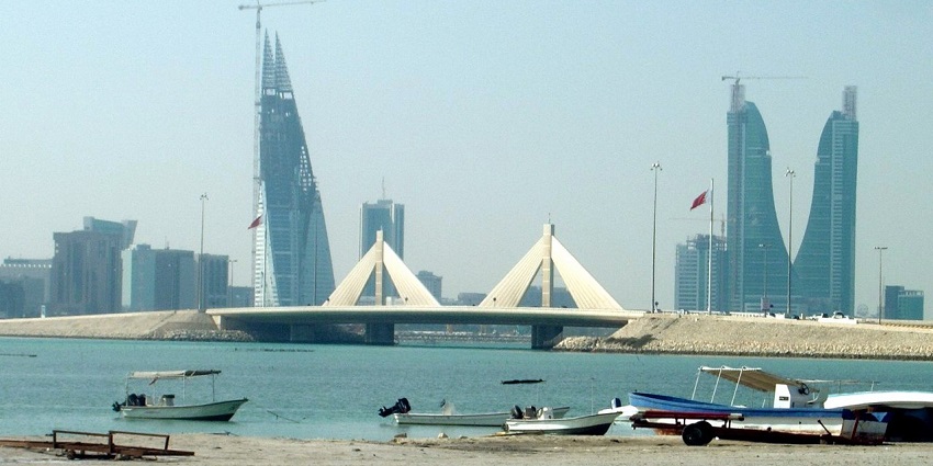 A view of the Bahrain skyline with modern skyscrapers, traditional boats on the water, and a clear blue sky.