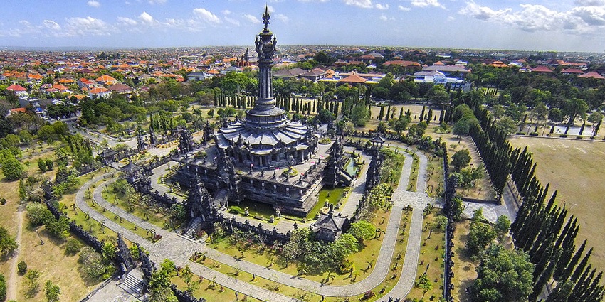 An aerial view of a Balinese-style structure with stone carvings, a central towering spire, and surrounding gardens and walkways.