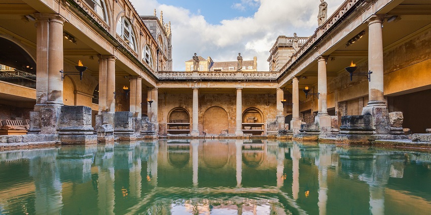 Roman baths with a clear water body of the bath, multiple pillars and fire torches on the wall.
