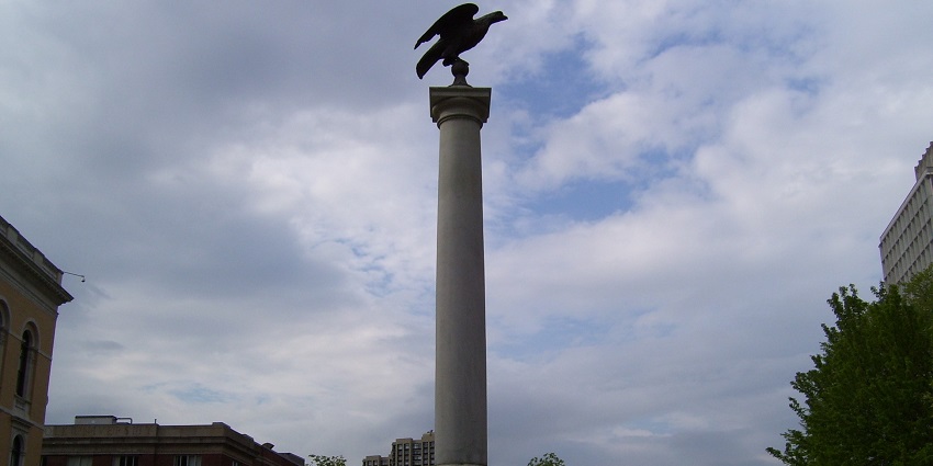 Column monument with eagle statue on top, set in a cityscape with trees and buildings.