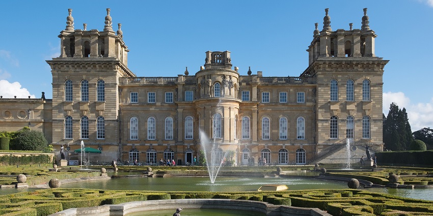 A view of Blenheim Palace with multiple fountains, statues near fountains and distant people.