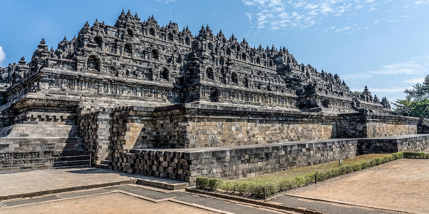 A massive ancient stone temple with intricate carvings and multiple tiers stands under a bright blue sky with scattered clouds.
