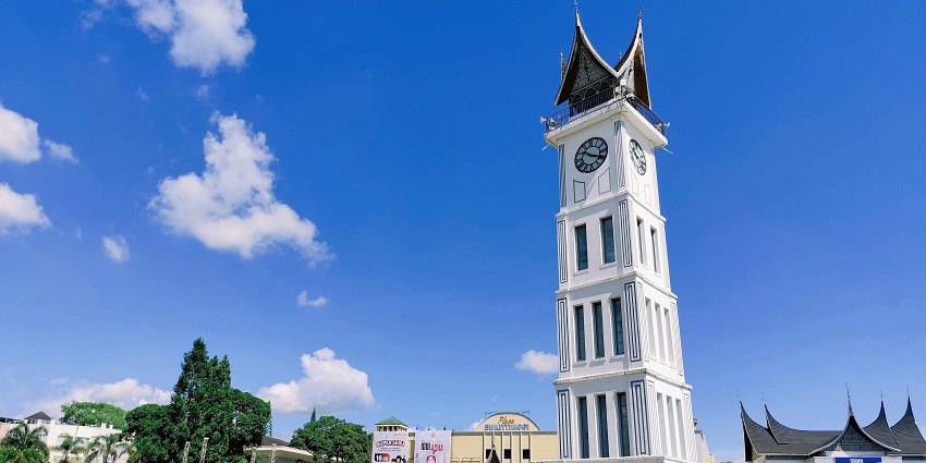 A tall clock tower stands in a busy town square with a mountain backdrop.