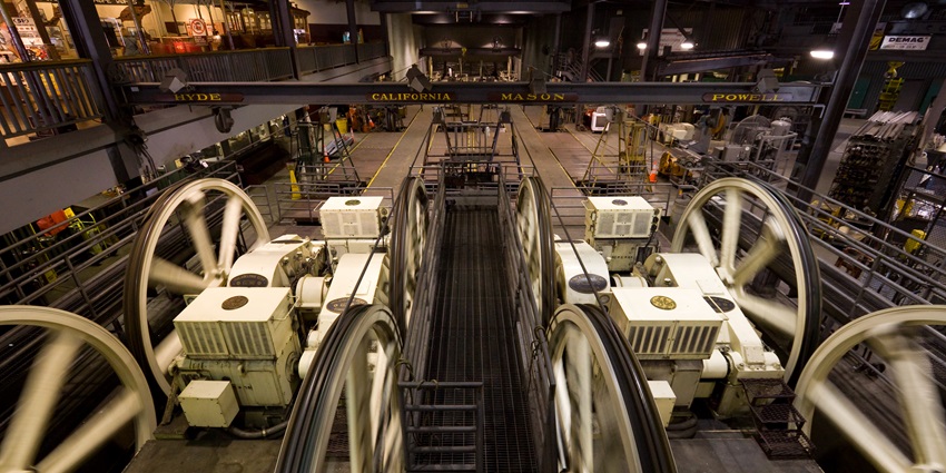Large mechanical wheels and machinery inside a cable car powerhouse with route names.