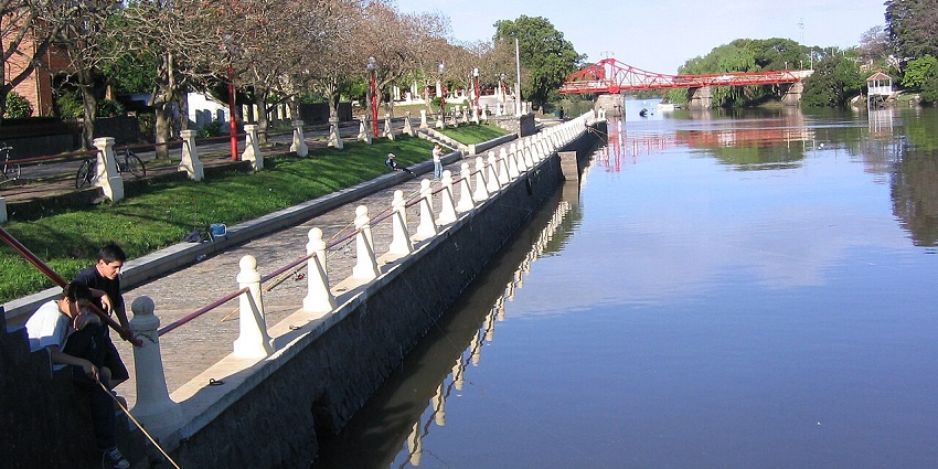A peaceful riverside promenade with people fishing and a red bridge in the background is a relaxing place to visit in Uruguay.

