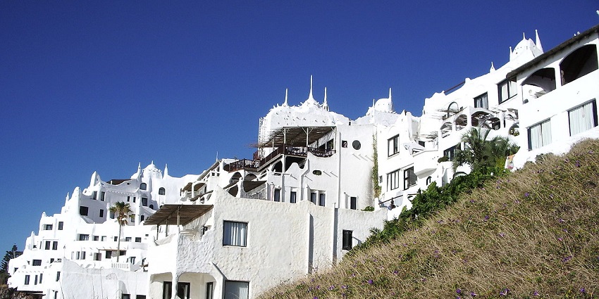 A cluster of white, irregularly shaped buildings with organic architecture clings to a hillside under a clear blue sky.