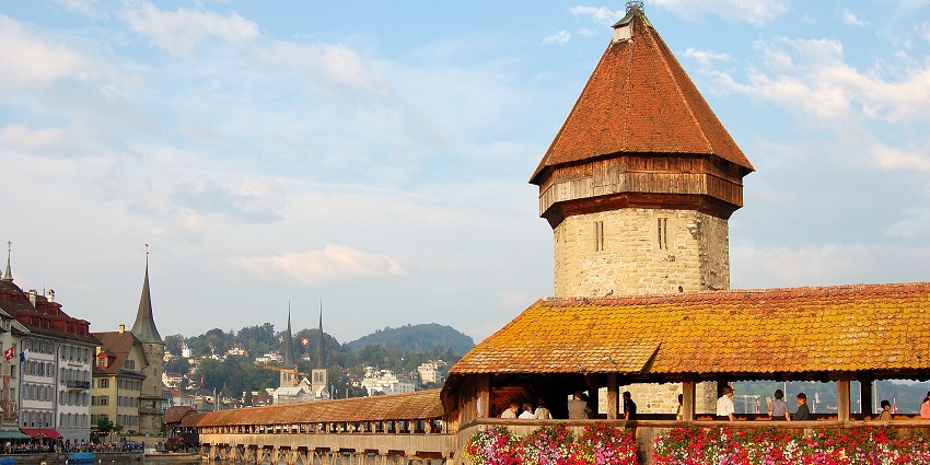 The Kapellbrücke wooden bridge in Lucerne, Switzerland, crossing the Reuss River with historic buildings and mountains in the background.