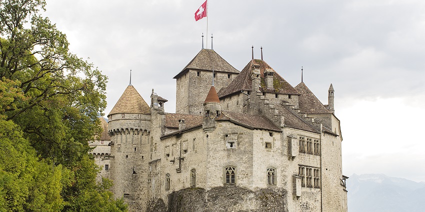 Château de Chillon castle beside Lake Geneva, surrounded by greenery under a cloudy sky, one of the top monuments in Switzerland.