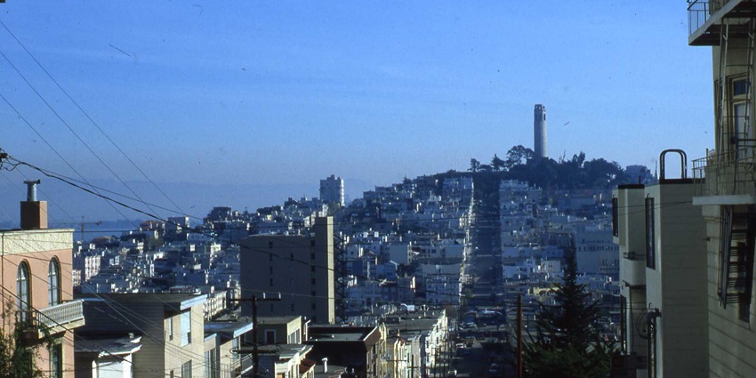 A steep city street lined with buildings leads to a distant tower on a hill under the sky.