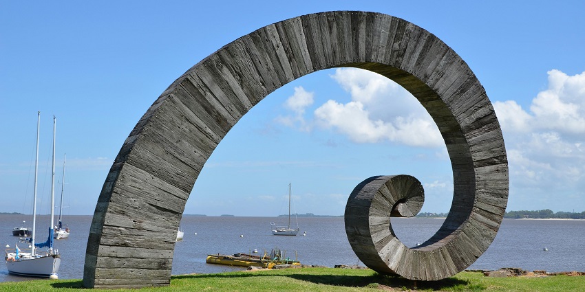 Large spiral wooden sculpture on a grassy waterfront with sailboats anchored in the calm water under a clear blue sky.