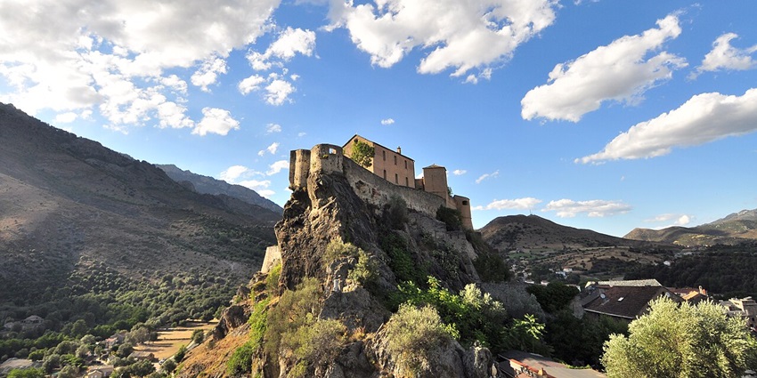 View of Corte Citadel with buildings on the rocky cliff and surrounded by green mountains.