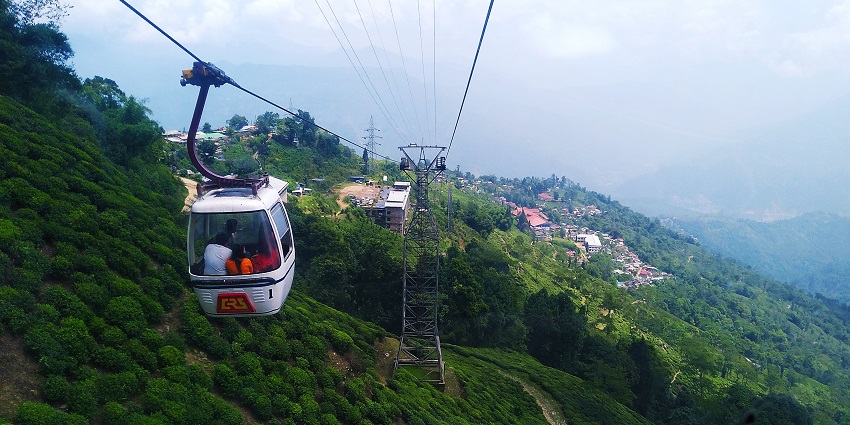 Darjeeling ropeway with people inside a white ropeway trolley, a tower and distant buildings.