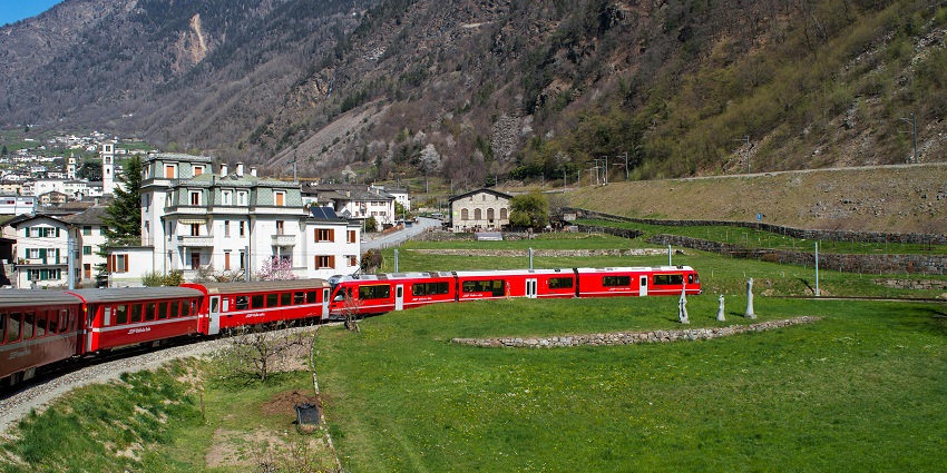 A bright red train winds through a scenic mountain village surrounded by lush green fields