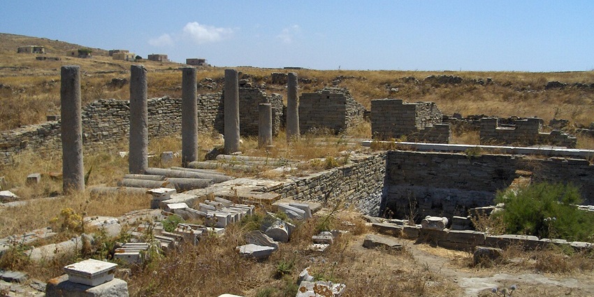 Ruins of ancient Greek structures with stone columns and walls in a dry, grassy landscape.