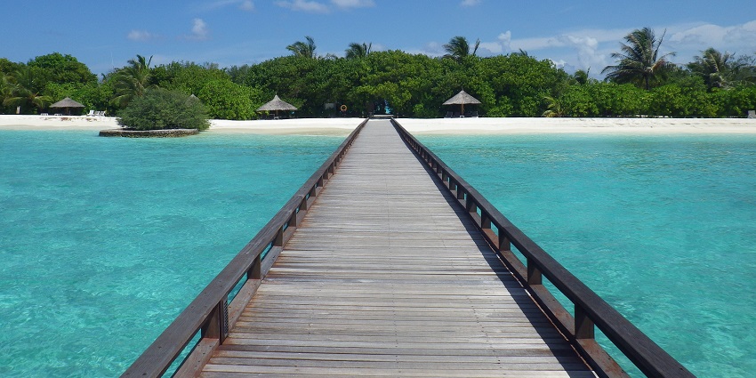 A wooden pier stretches over crystal-clear turquoise water toward a white sandy beach lined with palm trees and thatched huts.