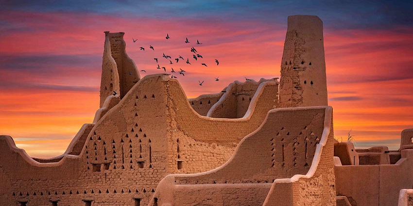 Al Turaif district with sandstone old buildings and multiple birds flying against a colourful sky
