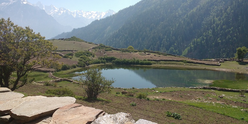 Serene mountain pond surrounded by lush greenery, stone fences, and distant snow peaks, one of the things to do in Lahaul and Spiti.