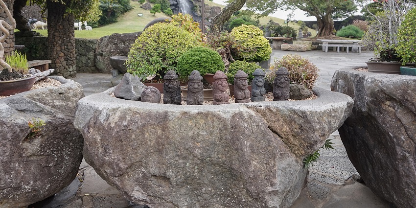 Stone statues called Dol Hareubangs stand in Jeju Stone Park, surrounded by trees and grass on a clear day.