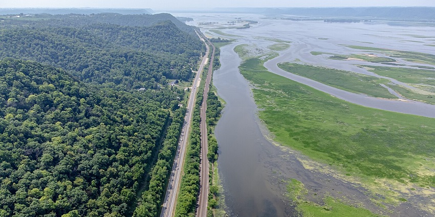 Scenic aerial view of the Great River Road with winding pavement, green trees, and Mississippi River visible alongside the route.