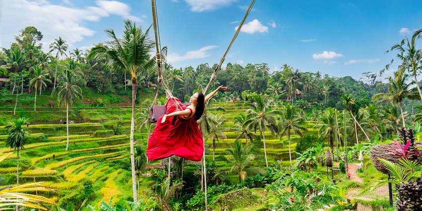Happy young female tourist in red dress enjoying the Bali swing at tegalalang rice terrace in Bali