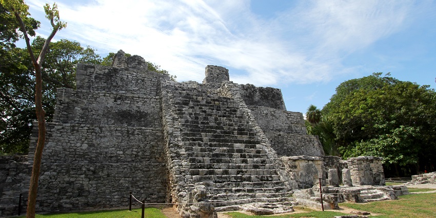 Ancient stone pyramid ruins with a central staircase, surrounded by green grass and trees.