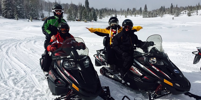 A group of people on a snowmobile tour, standing on a snowy mountain slope with snowmobiles and scenic views.