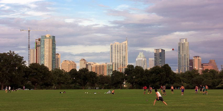 Wide grassy fields at Zilker Park with walking paths, large trees, and the distant downtown skyline.