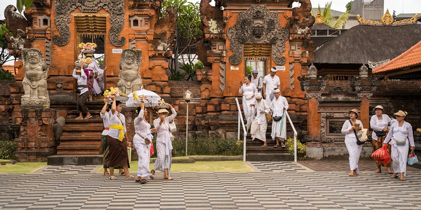 Devotees in traditional white attire gather before an ornate temple entrance, carrying offerings beneath ancient stone