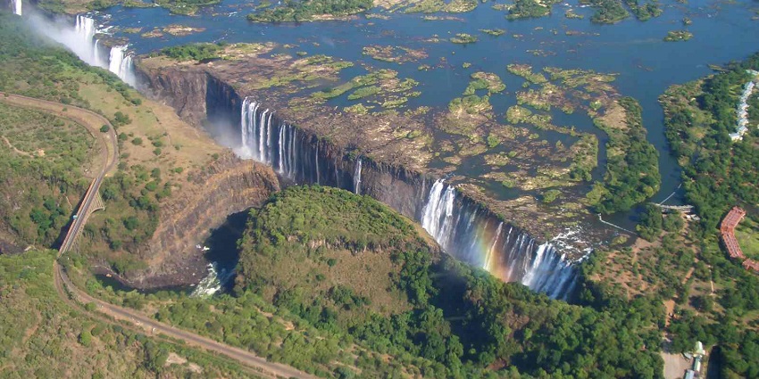 Tall waterfall with a visible rainbow under clear skies, crashing down into a rocky canyon below.