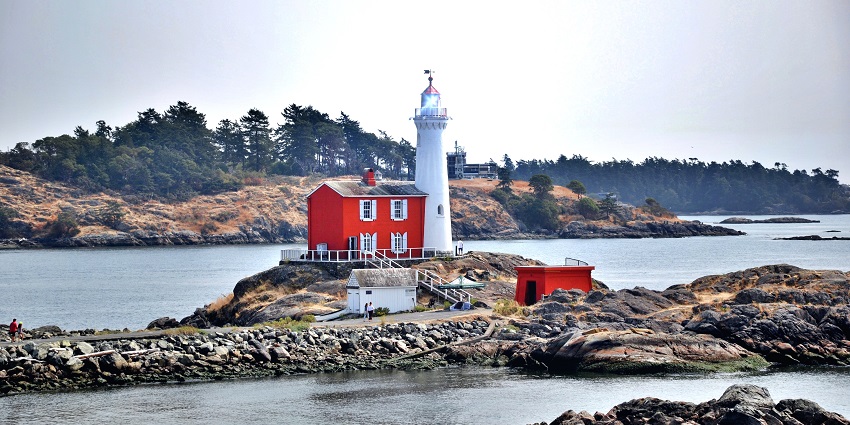 White lighthouse and red keeper’s house on a rocky shore with ocean and forested hills.