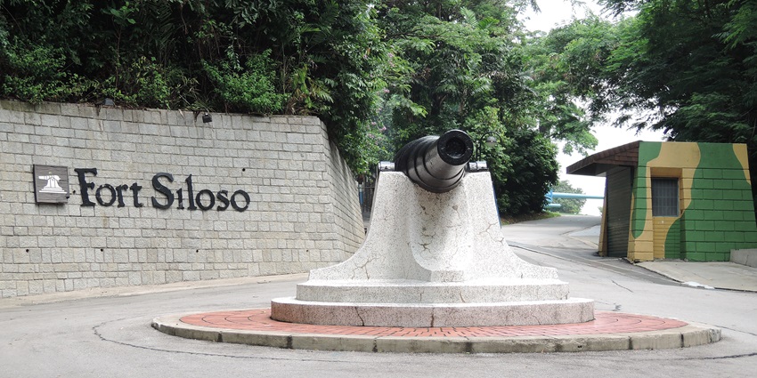 Entrance to Fort Siloso with a cannon statue, walkway, green trees, and a sign welcoming visitors to one of the historic places in Singapore.