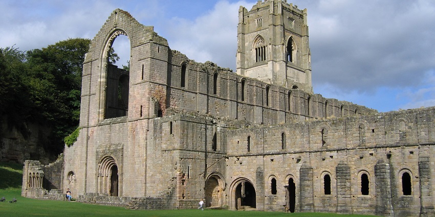 Ruins of Fountains Abbey in North Yorkshire with people visiting, broken walls and greenery.
