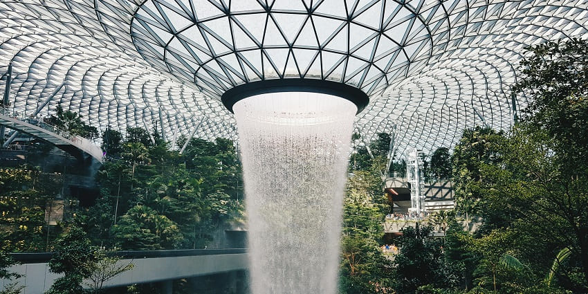 Indoor waterfall cascading from a glass-domed roof surrounded by lush green plants and trees.
