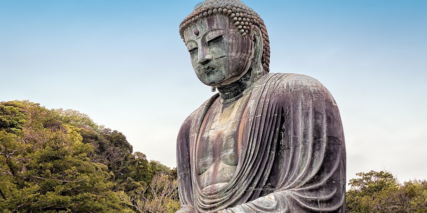 Large bronze Buddha statue seated outdoors, surrounded by trees under a clear sky.