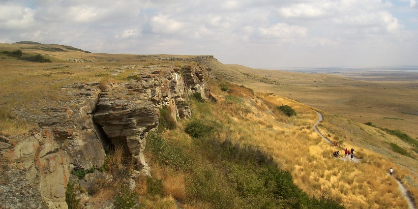 Head-Smashed-In Buffalo Jump with rocky cliffs, green plains and people on the small trail