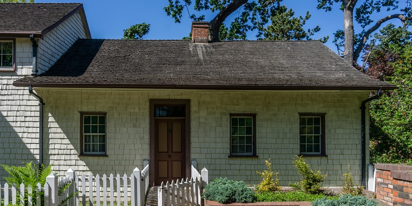 Cottage-style house with wood shingle walls, brown door, white picket fence, and garden.