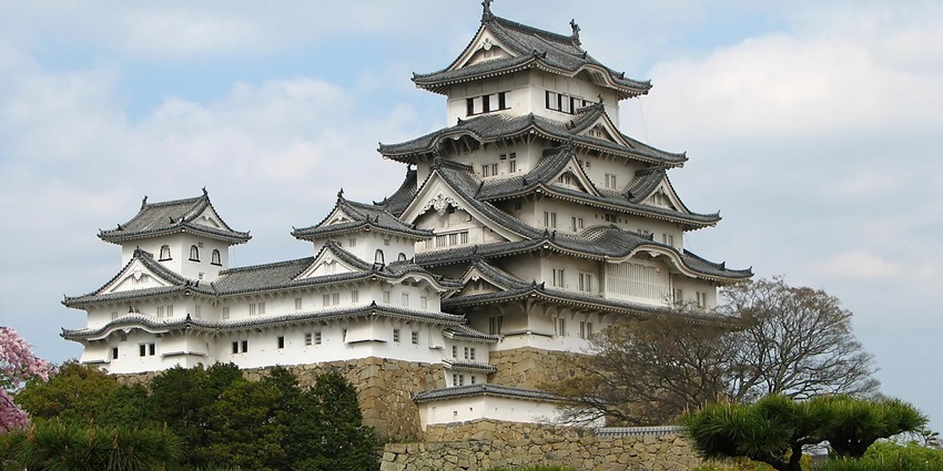 A traditional Japanese castle with layered roofs surrounded by trees under a cloudy sky, one of the historical places in Japan.