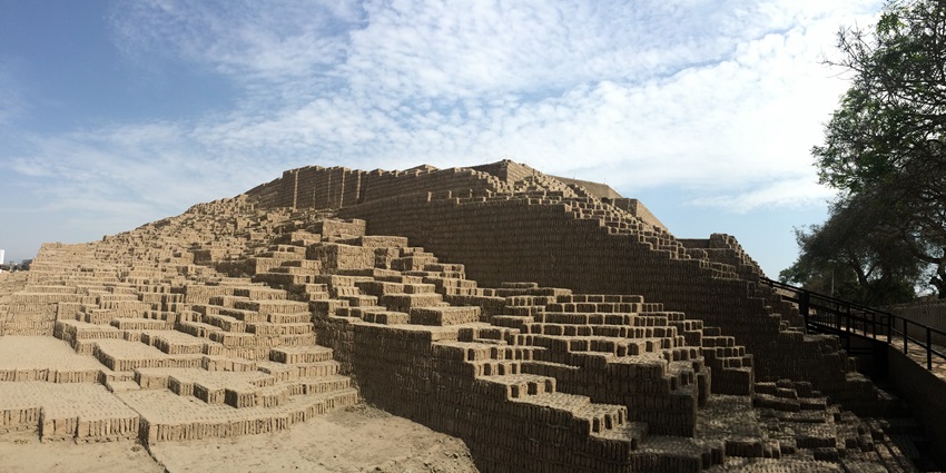 An ancient stepped pyramid made of sun-dried adobe bricks rises under a partly cloudy sky.