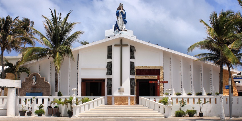 White church with a statue of the Virgin Mary, flanked by palm trees under blue sky.