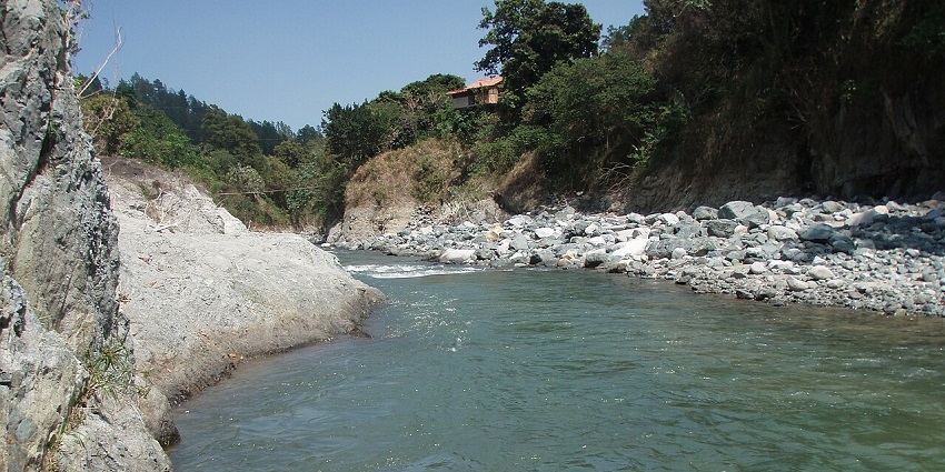 A clear river flows between rocky banks and tree-covered hills, with a small house perched above.