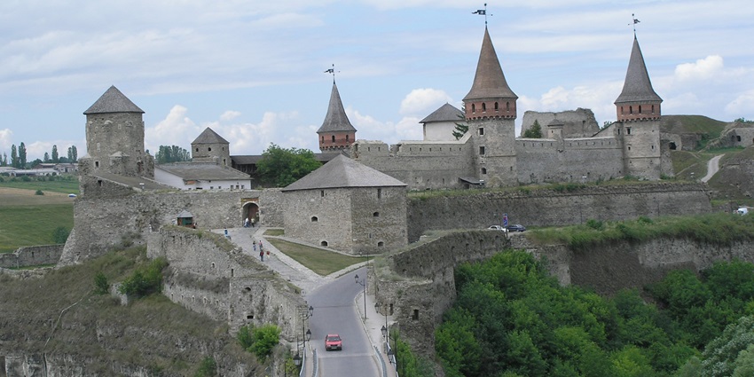 A medieval stone castle with towers and flags stands on a hill, connected by a bridge.