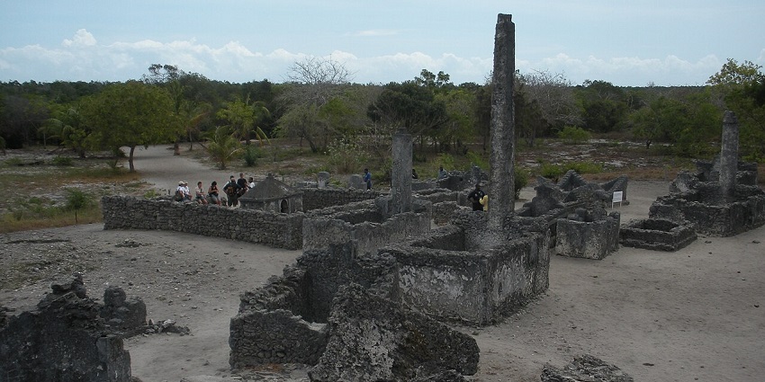 Ancient stone ruins of Kaole in Bagamoyo, Tanzania, surrounded by trees and greenery under a clear blue sky.
