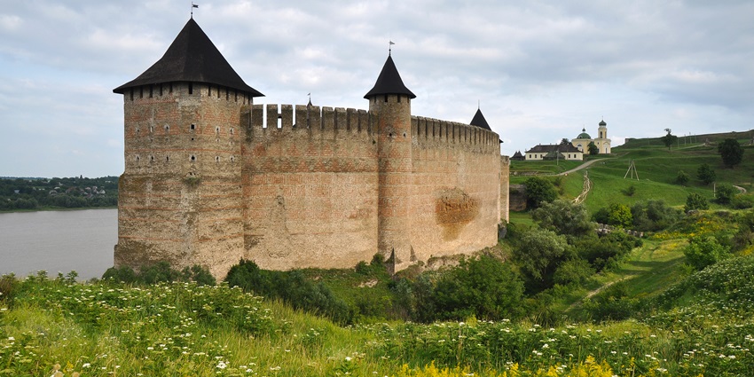 A large medieval fortress with conical towers beside a river, surrounded by green fields.