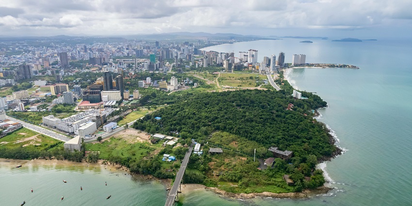 Aerial view of a coastal city in Cambodia with a lush green peninsula extending into the sea and high-rise buildings in the background.